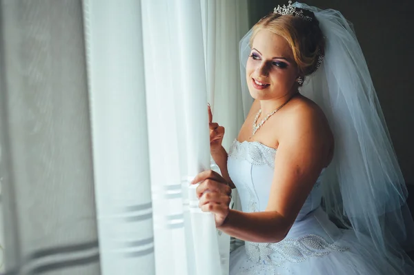 Beautiful young bride standing beside a large window waiting — Stock Photo, Image