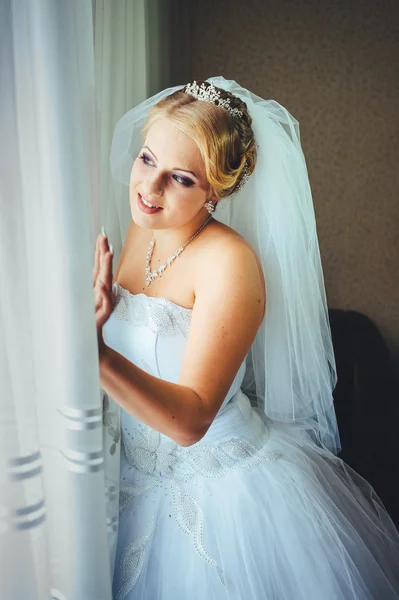 Beautiful young bride standing beside a large window waiting — Stock Photo, Image