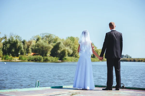 Um casamento à beira-mar. Lua-de-mel. A noiva e o noivo abraçados na margem do lago. noivo e noiva abraçando em um lago verde. O noivo e a noiva num parque. Vestido de noiva. Buquê de casamento nupcial de flores . — Fotografia de Stock