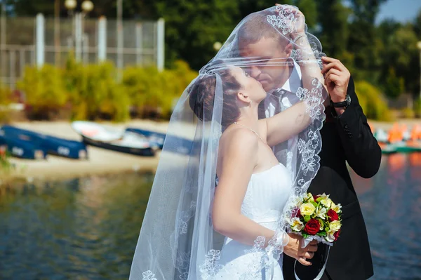 Um casamento à beira-mar. Lua-de-mel. A noiva e o noivo abraçados na margem do lago. noivo e noiva abraçando em um lago verde. O noivo e a noiva num parque. Vestido de noiva. Buquê de casamento nupcial de flores . — Fotografia de Stock