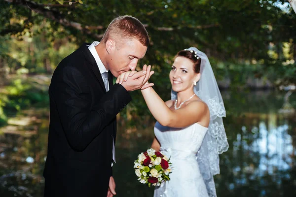 Um casamento à beira-mar. Lua-de-mel. A noiva e o noivo abraçados na margem do lago. noivo e noiva abraçando em um lago verde. O noivo e a noiva num parque. Vestido de noiva. Buquê de casamento nupcial de flores . — Fotografia de Stock