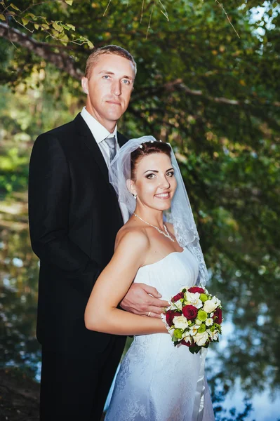 Um casamento à beira-mar. Lua-de-mel. A noiva e o noivo abraçados na margem do lago. noivo e noiva abraçando em um lago verde. O noivo e a noiva num parque. Vestido de noiva. Buquê de casamento nupcial de flores . — Fotografia de Stock