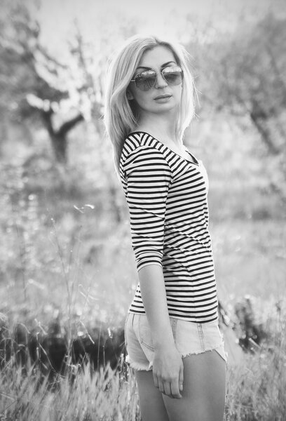 Blonde girl with glasses, shorts and sweater standing in the grass in black and white colors