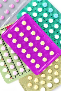 Temiz beyaz bir arkaplana yerleştirilmiş birden fazla renkli doğum kontrol hapı paketinin yüksek açılı yakın plan çekimi. Bu görüntü, kadın üreme sağlığı, aile planlaması, hormonal tedavi ve modern doğum kontrol yöntemlerini temsil etmektedir. İdeal