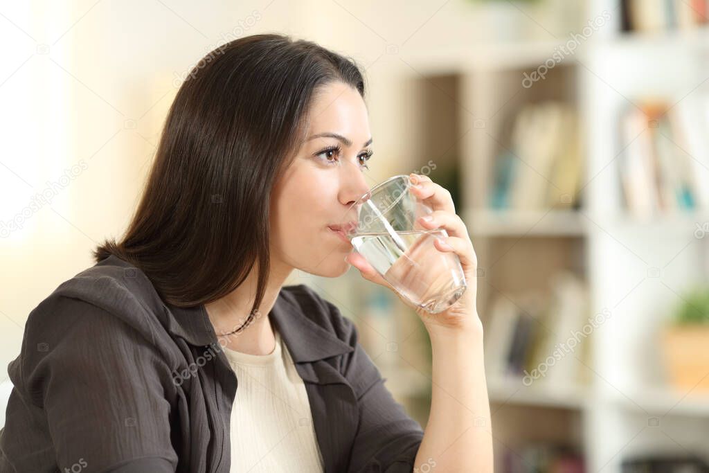 Woman drinking water from glass looking away at home
