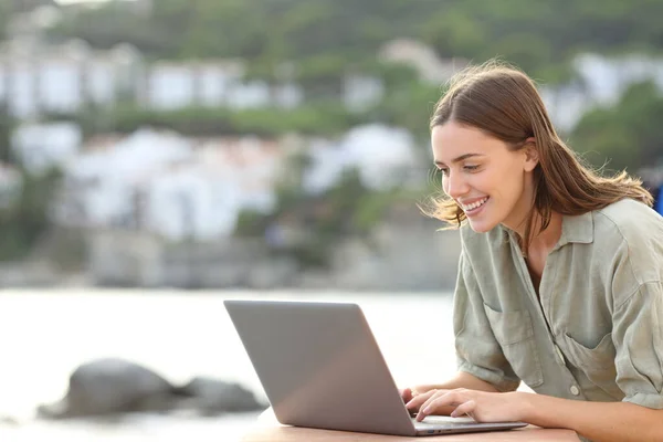 Happy woman writing on laptop in a balcony on the beach in summer