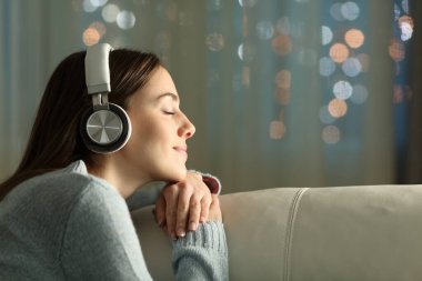 Gözü kapalı, kulaklıklı, kulaklıklı bir kadının profili gecenin bir yarısı evdeki bir kanepede oturup müzik dinliyor.