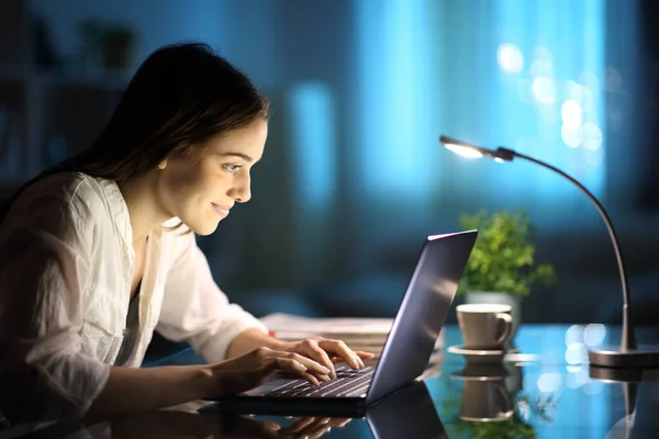 Happy woman writing on laptop keyboard on a desk in the night at home