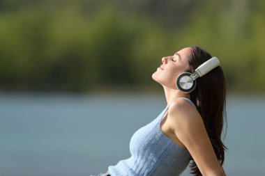Kulaklık takmış, gölde müzik dinleyen rahat bir kadının yan görüntüsü.