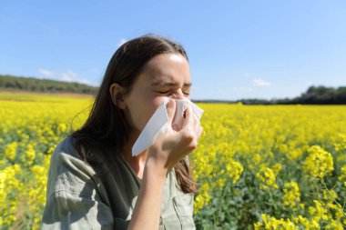 Allergic woman blowing nose standing in a field in spring season clipart