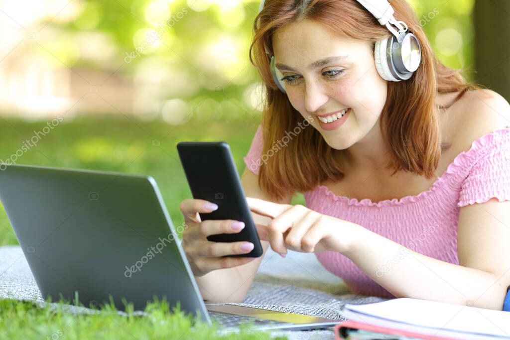 Happy student wearing headphones using laptop and smart phone in a park