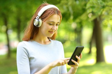 Kulaklık takan tatmin olmuş bir kadın parkta akıllı telefondan müzik dinliyor.