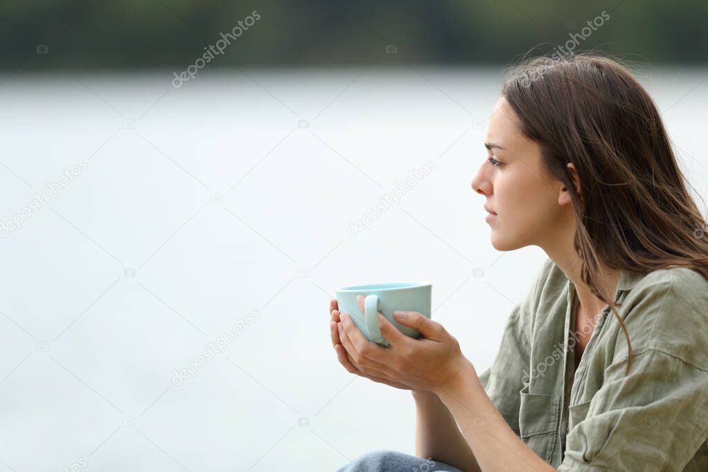 Side view portrait of a pensive woman holding coffee cup contemplating a lake