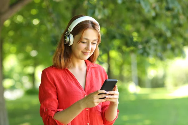 身穿红衫 头戴无线耳机的快乐女性正在公园里一边走路一边听智能手机上的音乐 — 图库照片