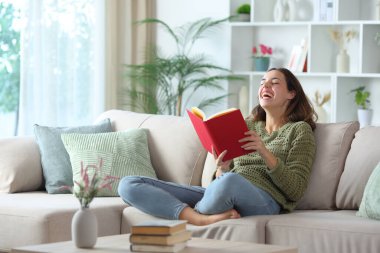 Mutlu kadın evde bir kanepede oturmuş komik bir komedi kitabı okurken gülüyor.
