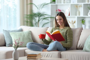 Yeşiller içindeki mutlu kadın kahve içiyor ve evdeki kanepede oturmuş gazete okuyor.