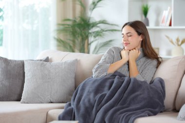 Evdeki kanepede battaniyeyle oturan gri elbiseli tatmin olmuş bir kadın.