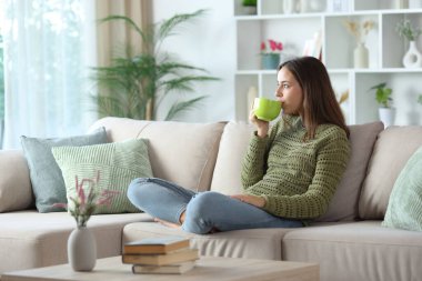 Yeşiller içinde kahve içen bir kadın evdeki kanepede oturuyor.