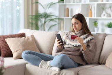 Kışın ciddi bir kadın internetten kredi kartı ve akıllı telefonuyla ev içinde bir kanepede oturuyor.