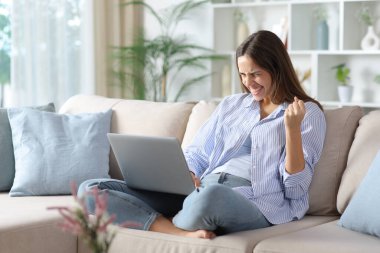 Mavili heyecanlı kadın iyi haberleri kutluyor evdeki koltukta dizüstü bilgisayarını kontrol ediyor.