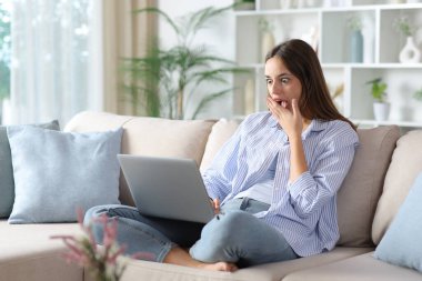 Mavili kadın evdeki kanepede dizüstü bilgisayarı kontrol ediyor.