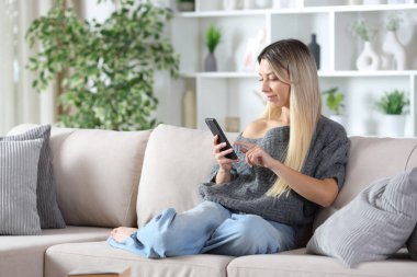 Sarışın kadın akıllı telefon kullanıyor. Evdeki koltukta oturuyor.