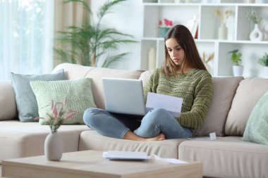 Evdeki bir kanepede oturup dizüstü bilgisayarını kullanan yeşil mektup yazan ciddi bir kadın.