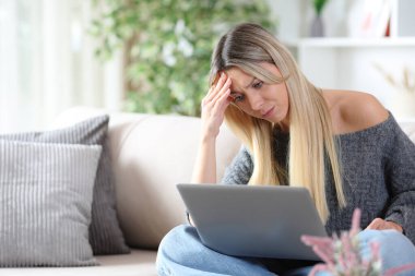 Endişeli kadın evdeki bir kanepede oturmuş kişisel bilgisayarından internet içeriğini kontrol ediyor.
