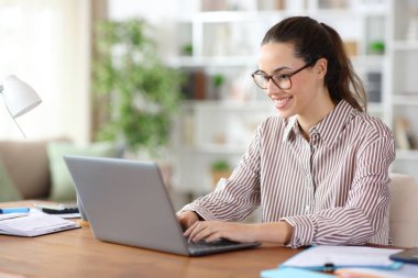 Evde dizüstü bilgisayar kullanarak çalışan gözlüklü mutlu bir tele-sekreter.