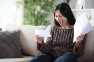 Heyecanlı Asyalı kadın çek ve mektup okuyor. Evdeki bir kanepede oturuyor.