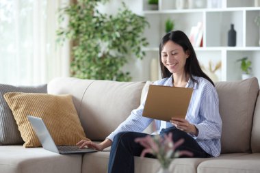 Mutlu Asyalı kadın evdeki kanepede oturan belgeleri ve laptopu kontrol ediyor.