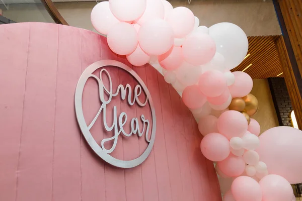 Beautiful festive decorations, pink and white balloons arch and number one on wooden round background. Little 1 year old girl birthday party photo zone.