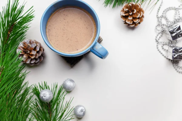 Blue cup of hot cacao or cappuccino with cinnamon standing on white table with pine branches and silver bulbs. Merry Christmas, Happy New Year and winter holidays concept. Copy space.