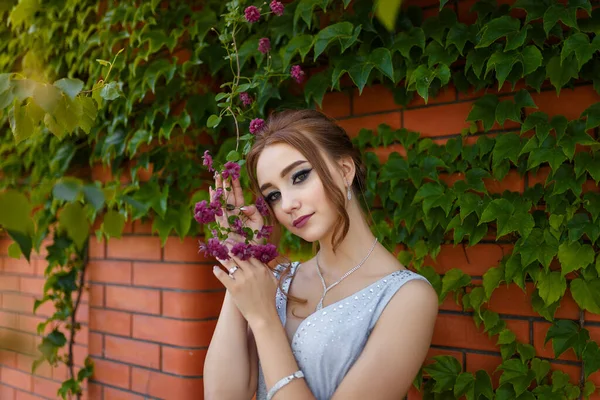 漂亮的姑娘穿着漂亮的舞会礼服 装饰着砖墙和绿色的常春藤树 背景是花朵 春景中的女性肖像 — 图库照片