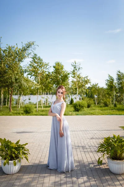 Beautiful girl in tender prom dress between two flowerbeds among green trees on blue sunny sky background. Female portrait on spring landscape.