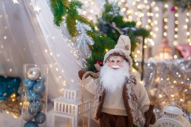 Kahverengi giyinmiş Noel Baba oyuncağı beyaz dekoratif fenerin önünde duruyor. Yeni yıl dekorasyonu, yılbaşı ışıl ışıl ışıkları ve odaklanmamış arka planda bokeh. Kış tatili sahnesi, mucize bekliyor..