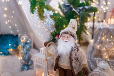 Kahverengi giyinmiş Noel Baba oyuncağı beyaz dekoratif fenerin önünde duruyor. Yeni yıl dekorasyonu, yılbaşı ışıl ışıl ışıkları ve odaklanmamış arka planda bokeh. Kış tatili sahnesi, mucize bekliyor..