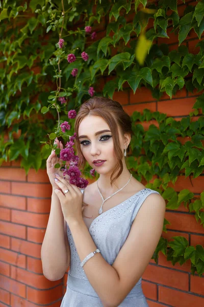 漂亮的姑娘穿着漂亮的舞会礼服 装饰着砖墙和绿色的常春藤树 背景是花朵 春景中的女性肖像 — 图库照片