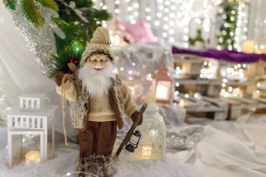 Kahverengi giyinmiş Noel Baba oyuncağı beyaz dekoratif fenerin önünde duruyor. Yeni yıl dekorasyonu, yılbaşı ışıl ışıl ışıkları ve odaklanmamış arka planda bokeh. Kış tatili sahnesi, mucize bekliyor..