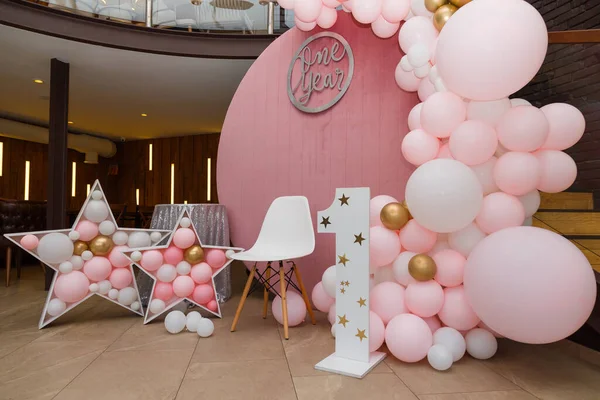 Beautiful festive decorations, pink and golden balloons arch, wooden stars, white chair and number one on wooden round background. Little 1 year old girl birthday party photo zone.