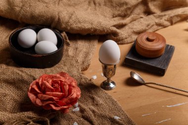 Yumurta kabında haşlanmış yumurta ve siyah kase, gümüş kaşık, tuz kabı ve çuval bezinde gül çiçeği ve ahşap kahverengi arka plan. Bahar ve Paskalya konsepti.