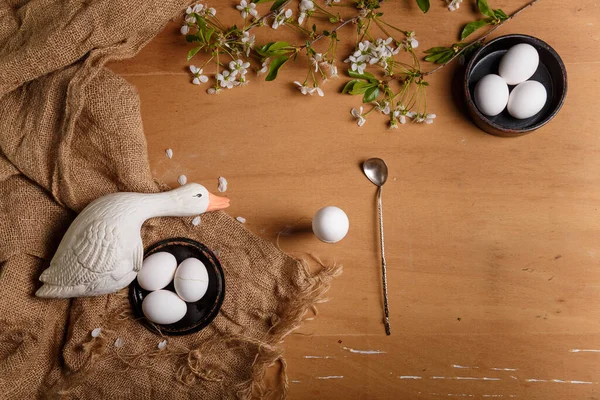 Easter eggs in black bowls, goose toy, spoon, boiled egg in egg cup, cherry blossoming branch with white flowers on sackcloth and brown wooden background. Top view, copy space.