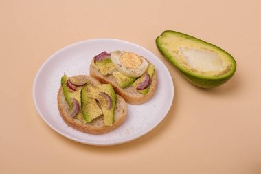 Avokado ile iki sandviç, haşlanmış yumurta ve beyaz tabakta kırmızı soğan, yarısı bej arka planda avokado. Asgari biçim kompozisyonu.
