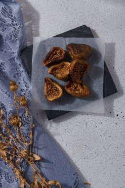 Kağıt üzerinde kurumuş incir parçaları, beyaz arka planda mavi kumaş ve kuru çiçekler. Dikey çekim, üst görüş. Sağlıklı beslenme, şekersiz.