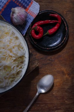 Bir kase lahana turşusu, sarımsak ve acı biber, mutfak havlusu ve tahta kırsal masa arka planında kaşık. Kış ve sonbahar için vejetaryen yemekleri.