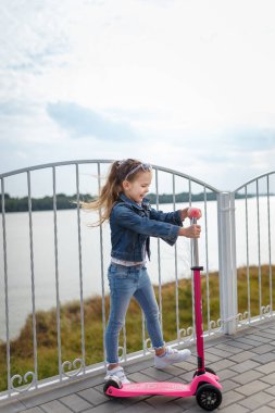 Tekme scooterlı, gülümseyen küçük kız. Dışarıda oynayan mutlu çocuk. Tasasız çocukluk.