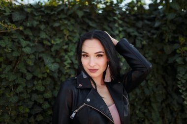 Siyah deri ceketli, yeşil sarmaşık arka planında poz veren güzel siyah saçlı bir kadının portresi..