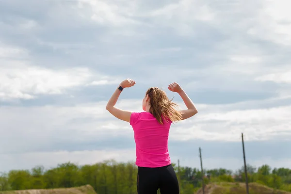 Young happy woman in sportswear standing on hill back to camera with arms up to sky. Fitness, healthy way of life, wellbeing, freedom, mental health, triumph, winning concept.