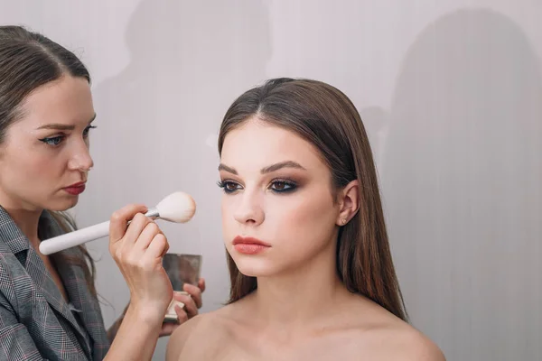 Make-up artist doing visage for beautiful teenage girl, using foundation for face and blush for cheeks. Make up, cosmetics, beauty standards, skin care concept. Close up shot.