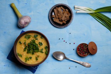 Bir kase bezelye ya da mercimek çorbası taze sarımsak, yeşil soğan, maydanoz, aromatik biber seti ve mavi ahşap arka planda çavdar ekmeği krakeri ile servis edilir. Sağlıklı beslenme. Üst görünüm, alanı kopyala.